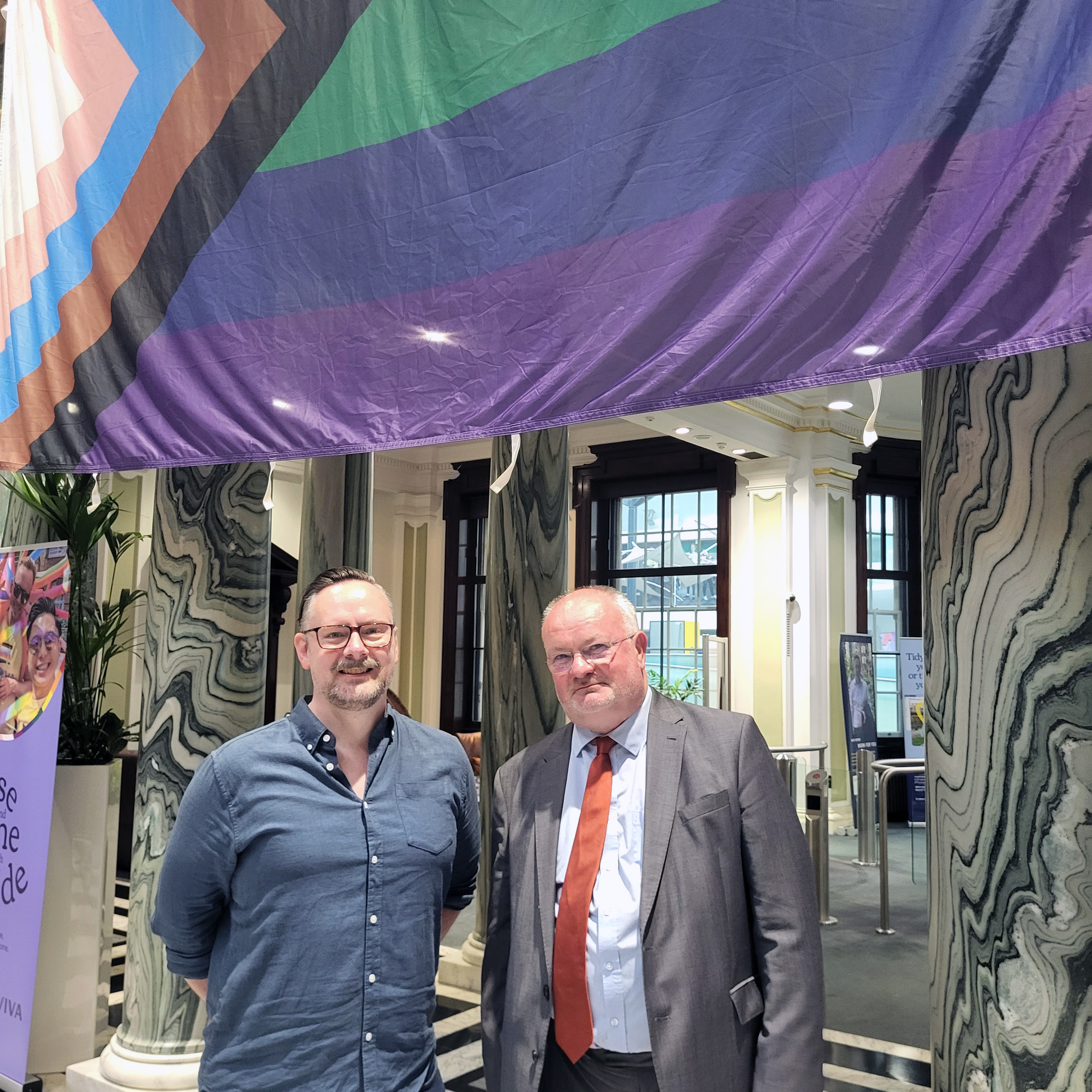 Robert George from Norwich Pride, and Piers Marlow from First Bus standing in front of a progress pride flag